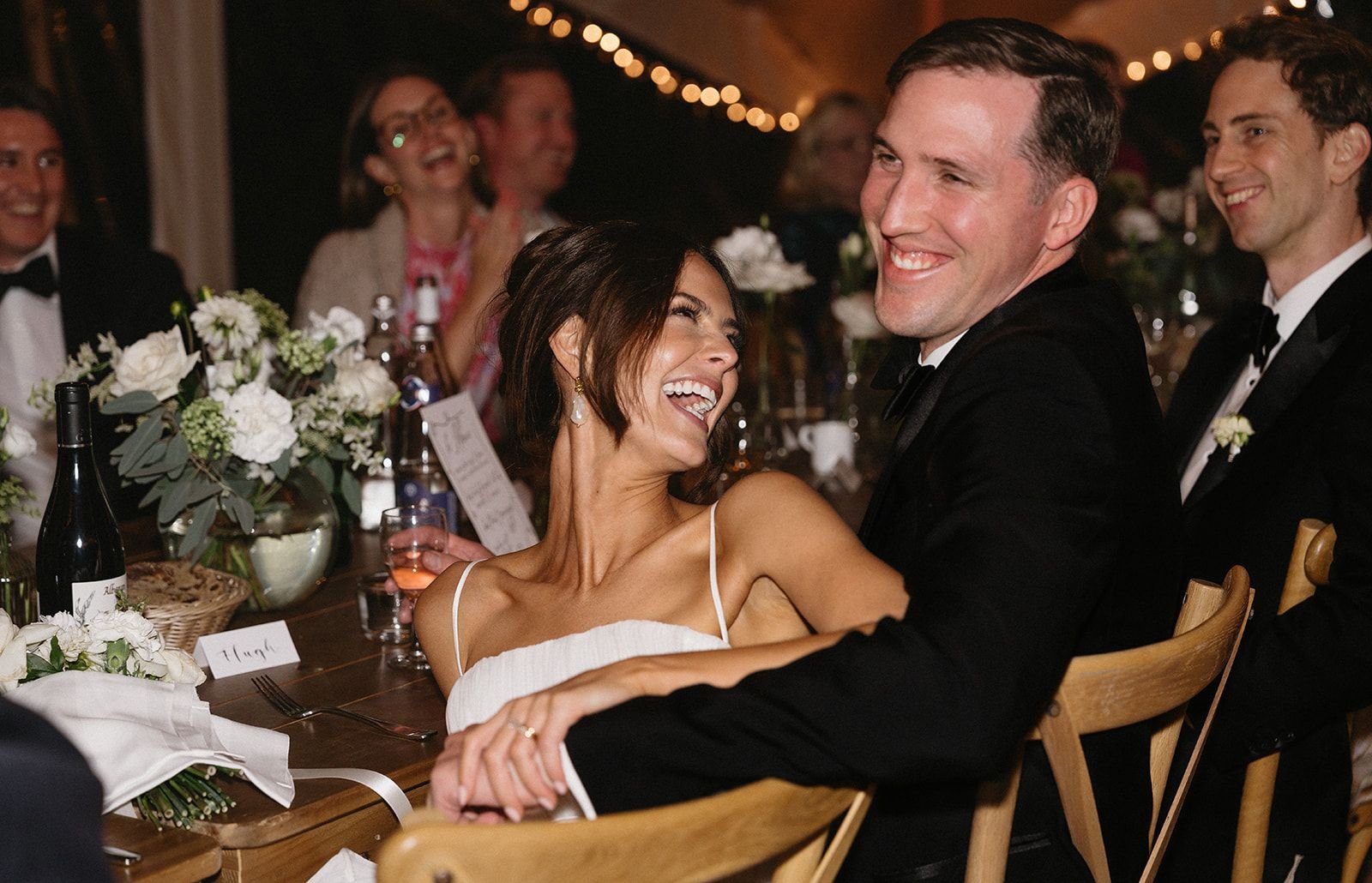 A woman in a white dress and a man in a black tuxedo are smiling and laughing together at a wedding reception. Other guests are visible in the background, also smiling and enjoying the celebration.