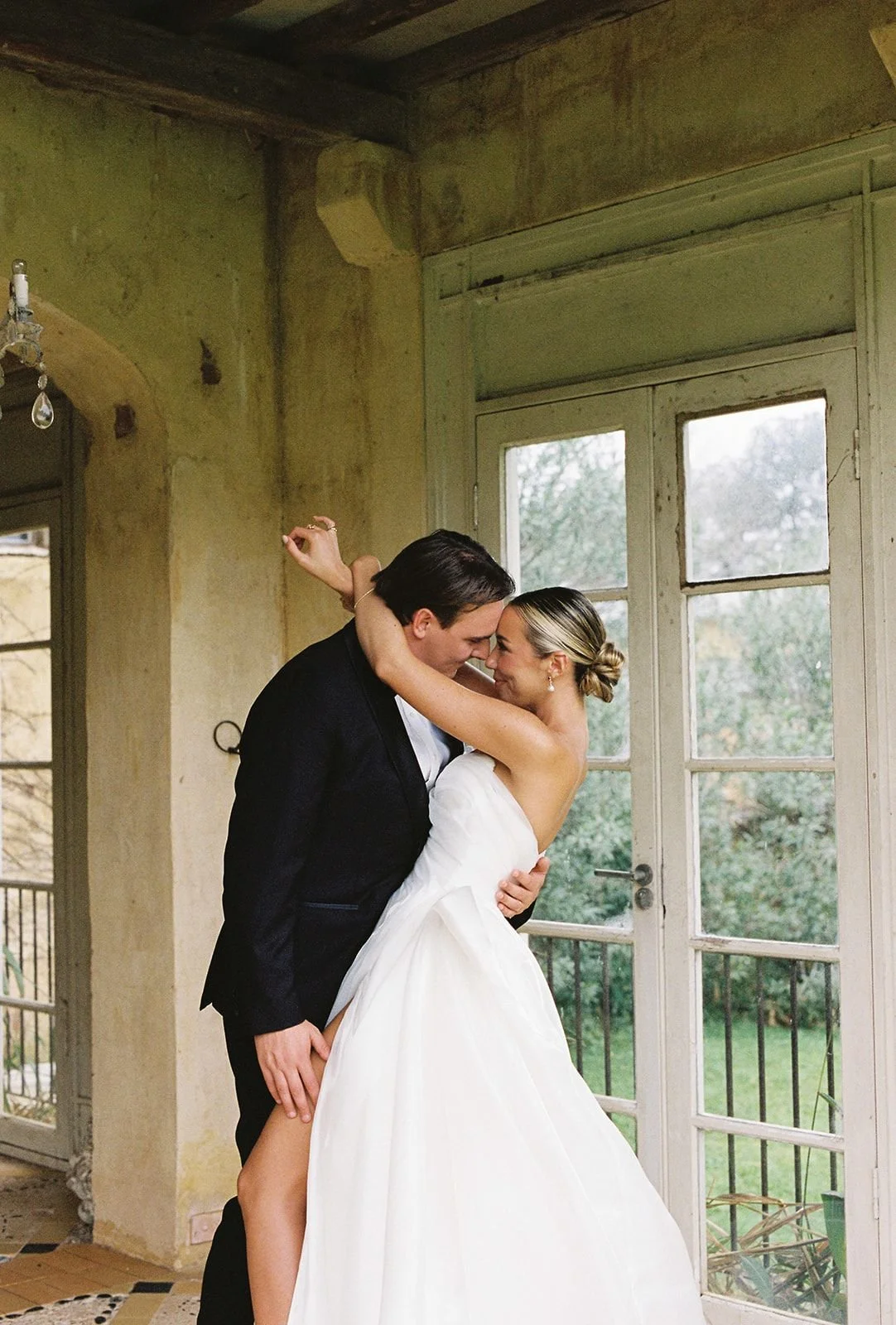 A bride and groom sharing a dance, standing close together indoors near large windows with greenery outside.