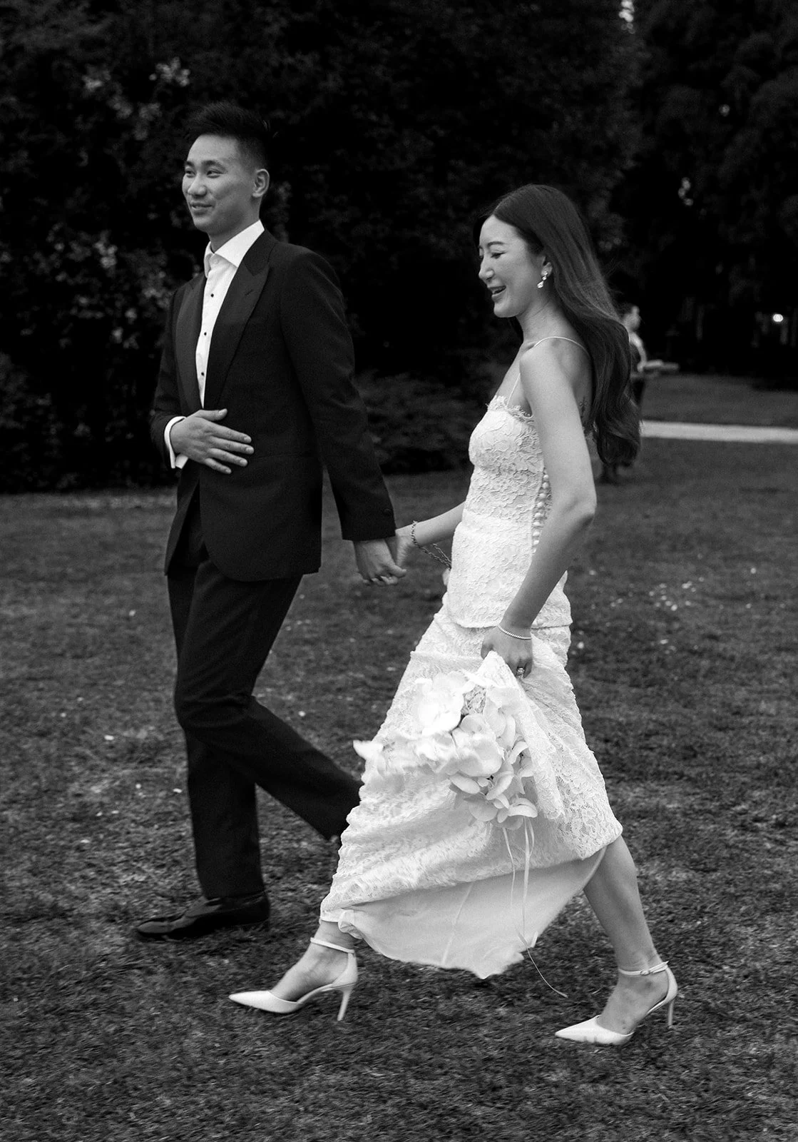 A black-and-white photo of a bride and groom holding hands and walking outdoors, the bride in a lace wedding dress and high heels, the groom in a tuxedo, surrounded by trees.