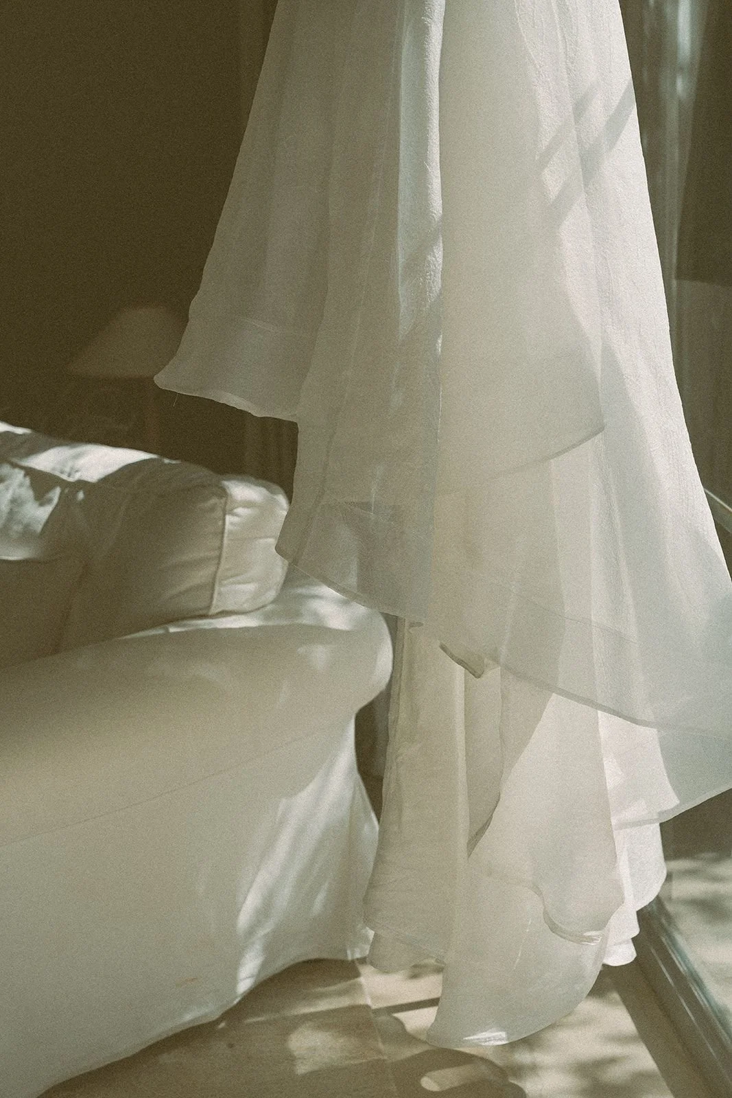 A sheer white curtain hanging in front of a window, with sunlight streaming through, casting shadows inside a room. Part of a white sofa is visible in the foreground.
