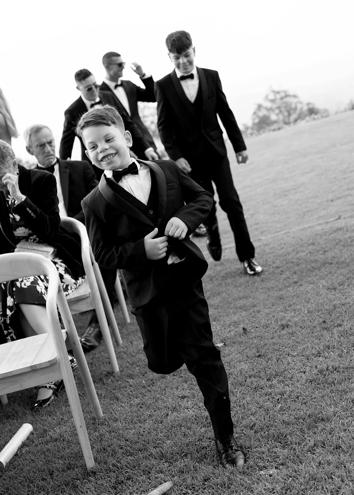 Young boys in tuxedos at a formal outdoor event, with one running happily and others walking in the background, some seated on chairs.