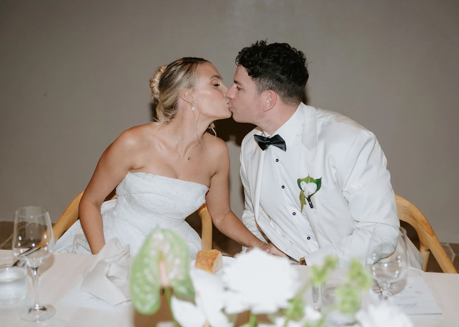 Bride and groom kiss at wedding reception table, wedding attire, floral decoration and table settings visible.