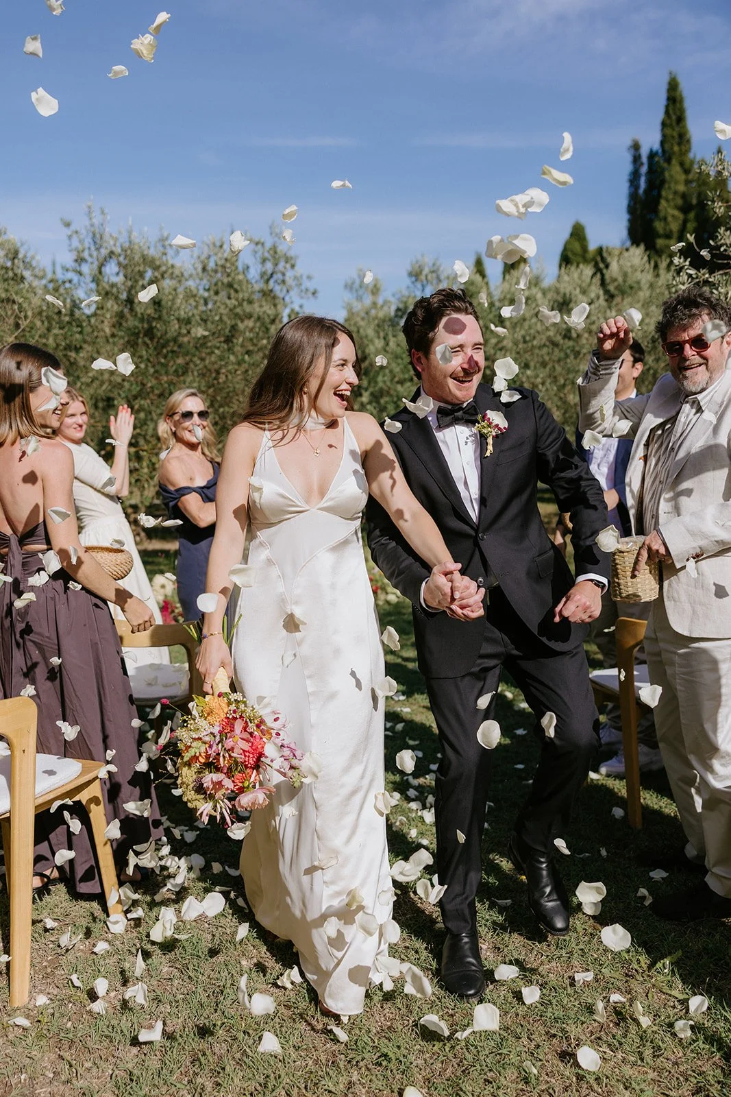 A newlywed couple holding hands and walking through falling flower petals at an outdoor wedding ceremony, surrounded by smiling guests on a sunny day.