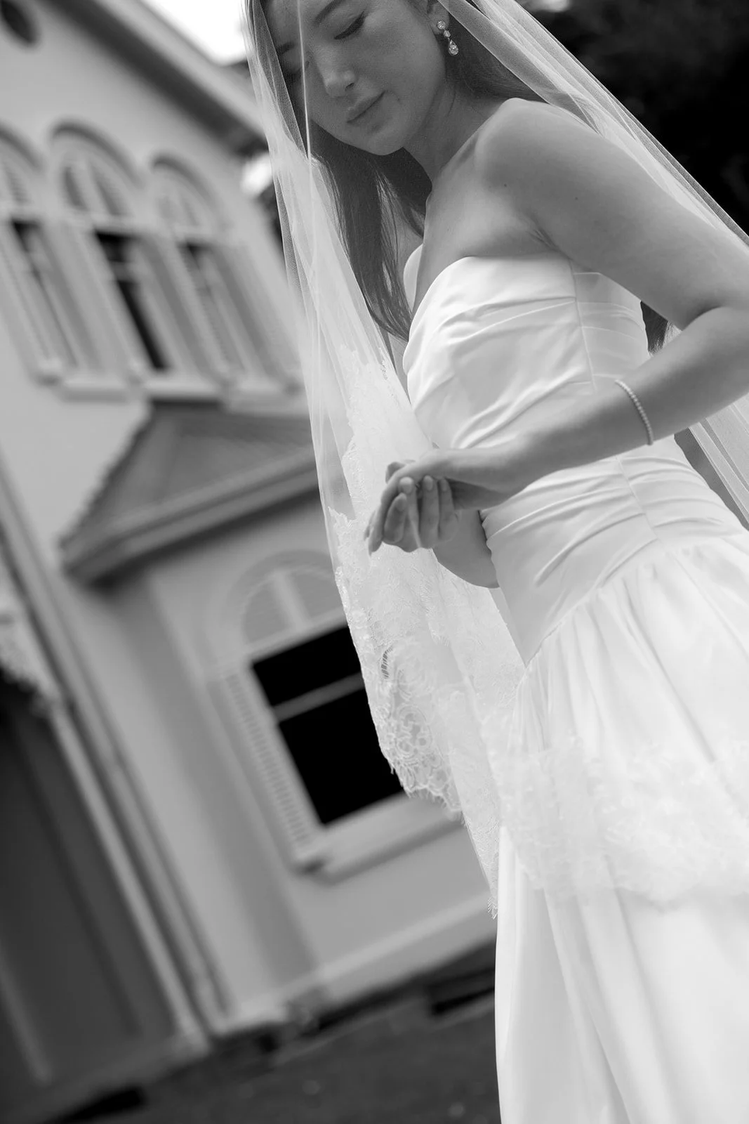 A bride wearing a strapless white wedding dress and veil, standing outside a building with arched windows, looking down thoughtfully.