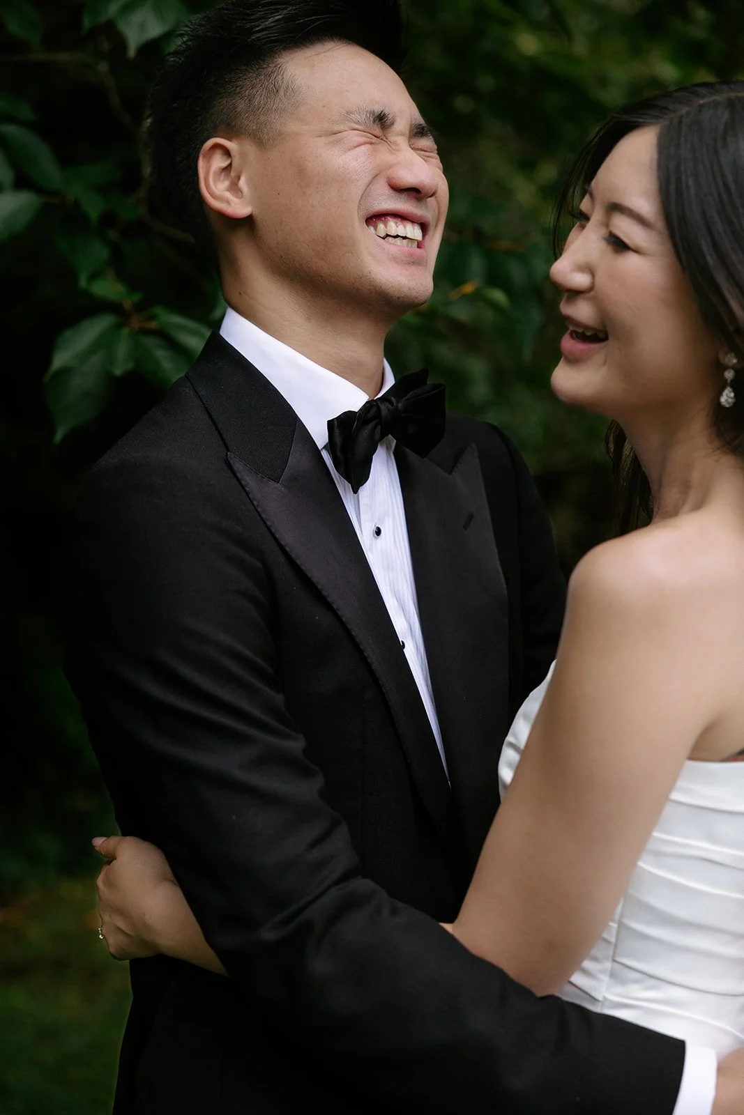 A happy bride and groom in wedding attire, embracing outdoors, with greenery in the background.