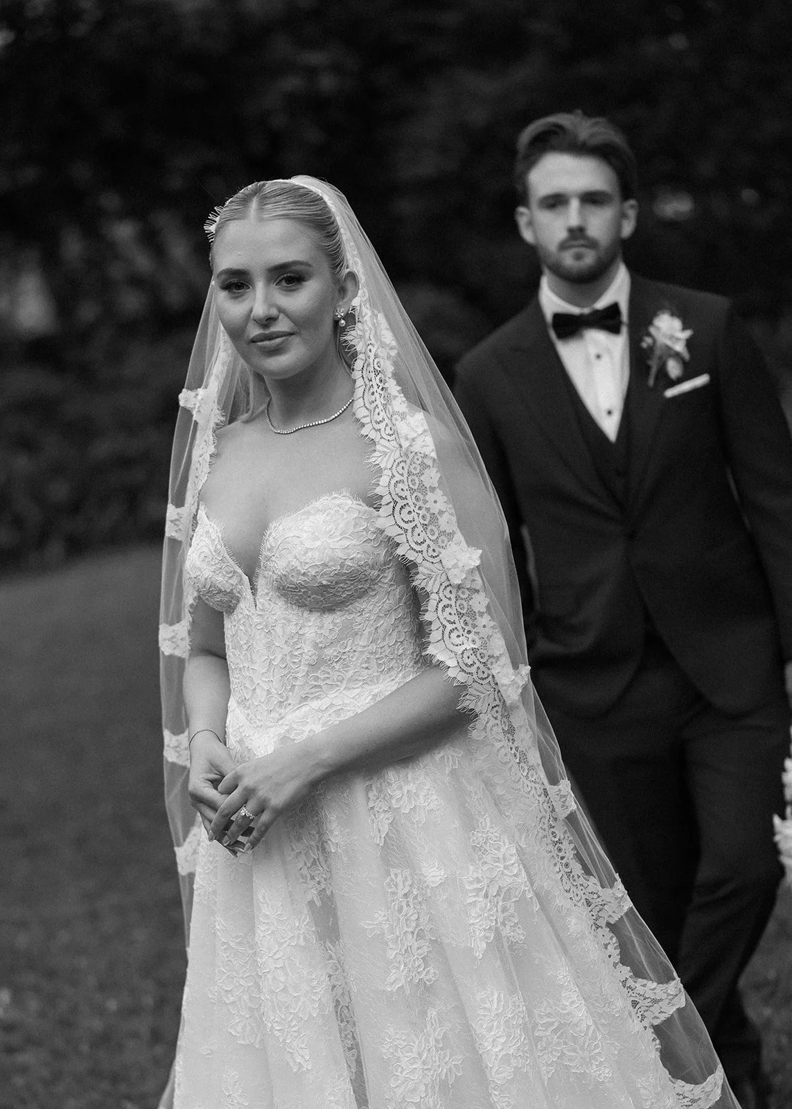 Black and white photo of a bride in a lace wedding dress with a veil, wearing jewelry, and a groom in a tuxedo with a bow tie and boutonniere, outdoors.