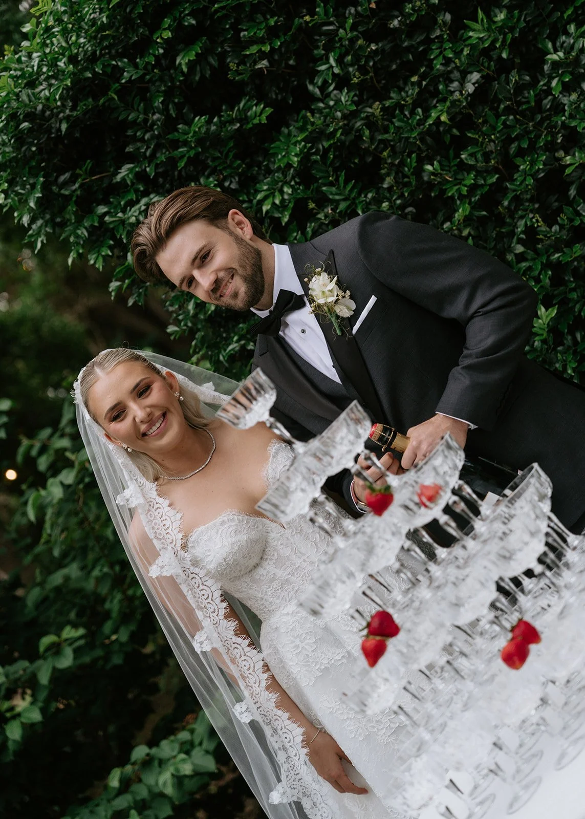 A newlywed couple in wedding attire smiling and standing outdoors next to a tiered champagne or cocktail glass display with strawberries, with green foliage in the background.
