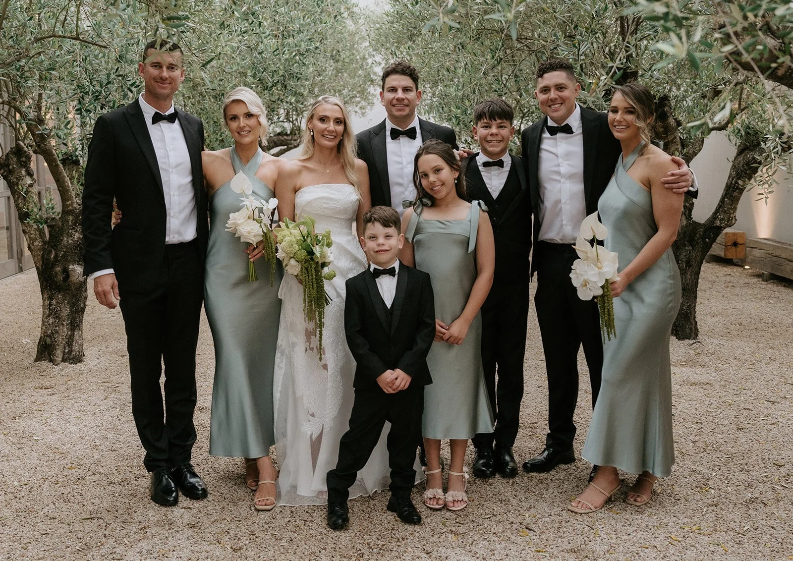 A group of people dressed in formal attire, including a bride in a white wedding dress holding a bouquet, and others in suits and dresses, standing outdoors on a gravel surface with trees in the background.