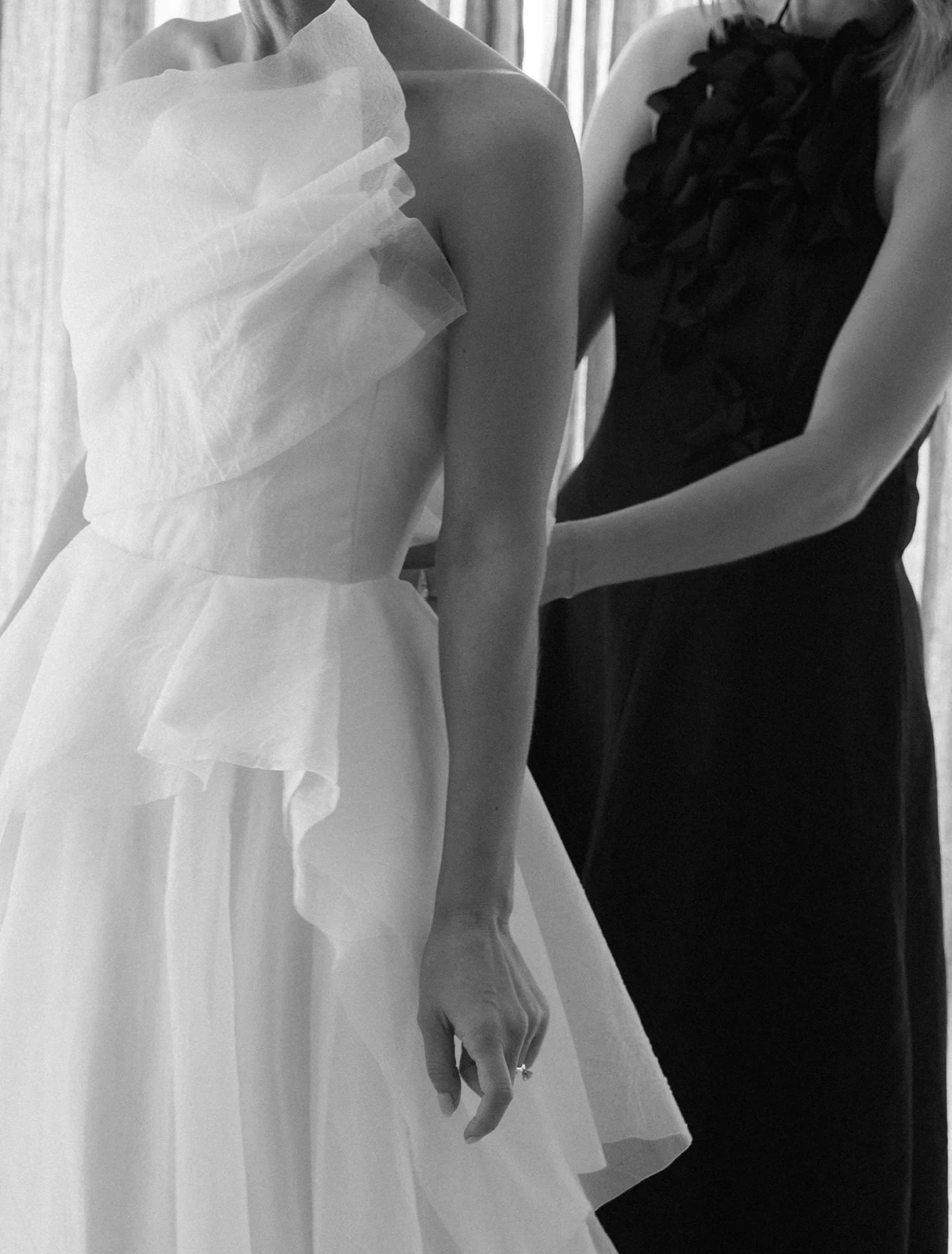 A woman in a wedding dress with ruffled tulle details is having the final adjustments made to her gown by another woman in a black sleeveless top with ruffles.