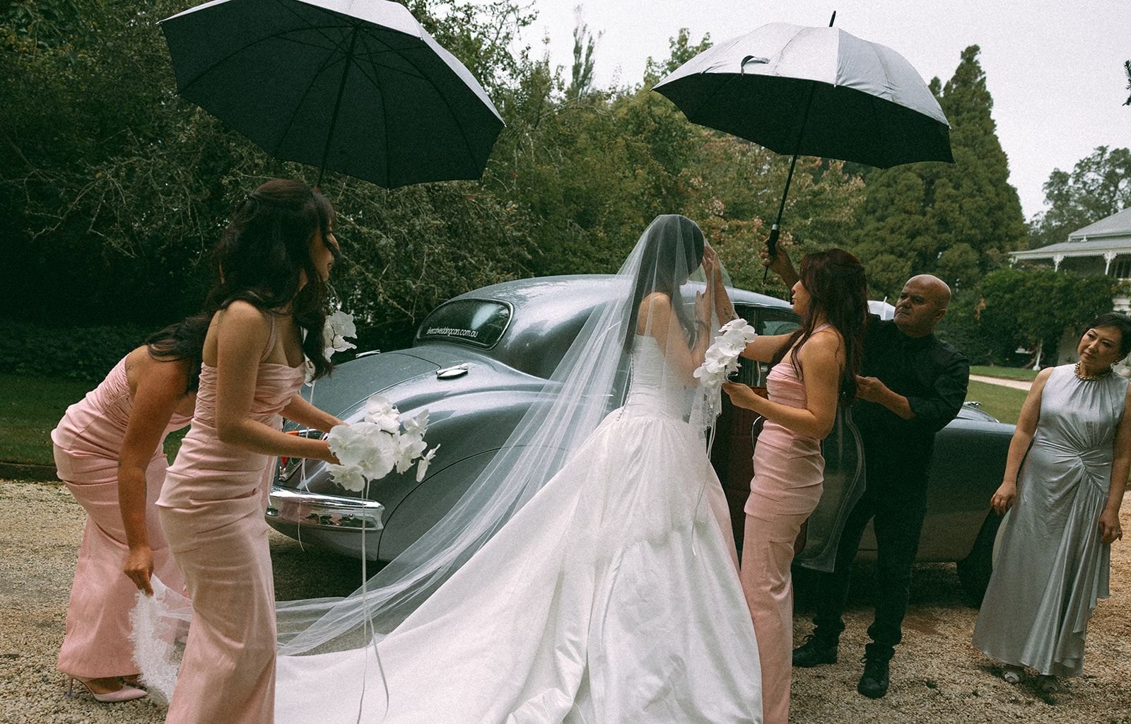 A bride in a wedding gown with a veil stands next to a vintage car as four women, dressed in pink dresses, hold umbrellas and flowers. A man in black assists her, and a woman in a silver dress looks on.