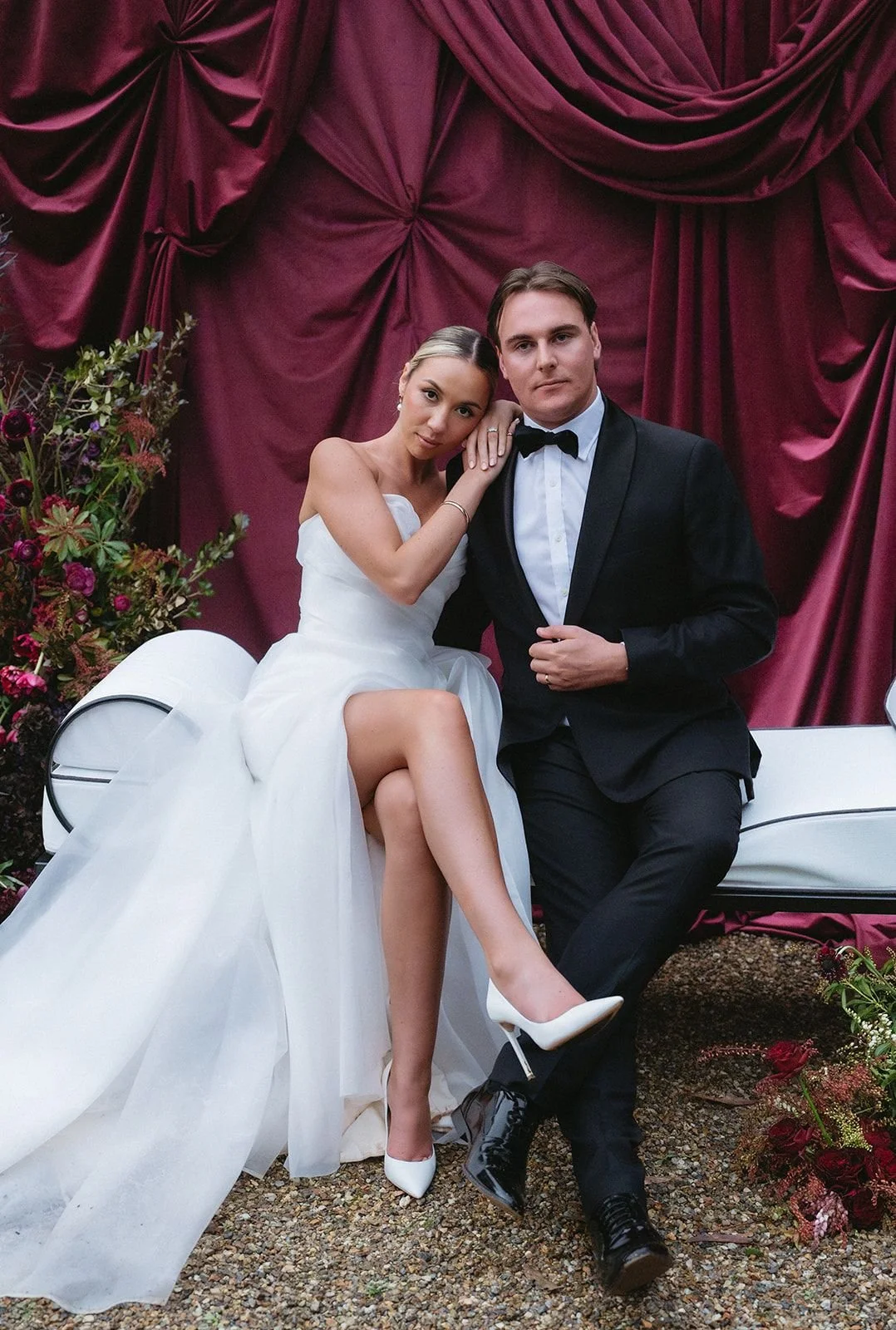 A bride and groom sitting on a white bench in front of a burgundy curtain, with the bride in a white strapless wedding gown and white heels, and the groom in a black tuxedo with a bowtie, surrounded by floral arrangements.