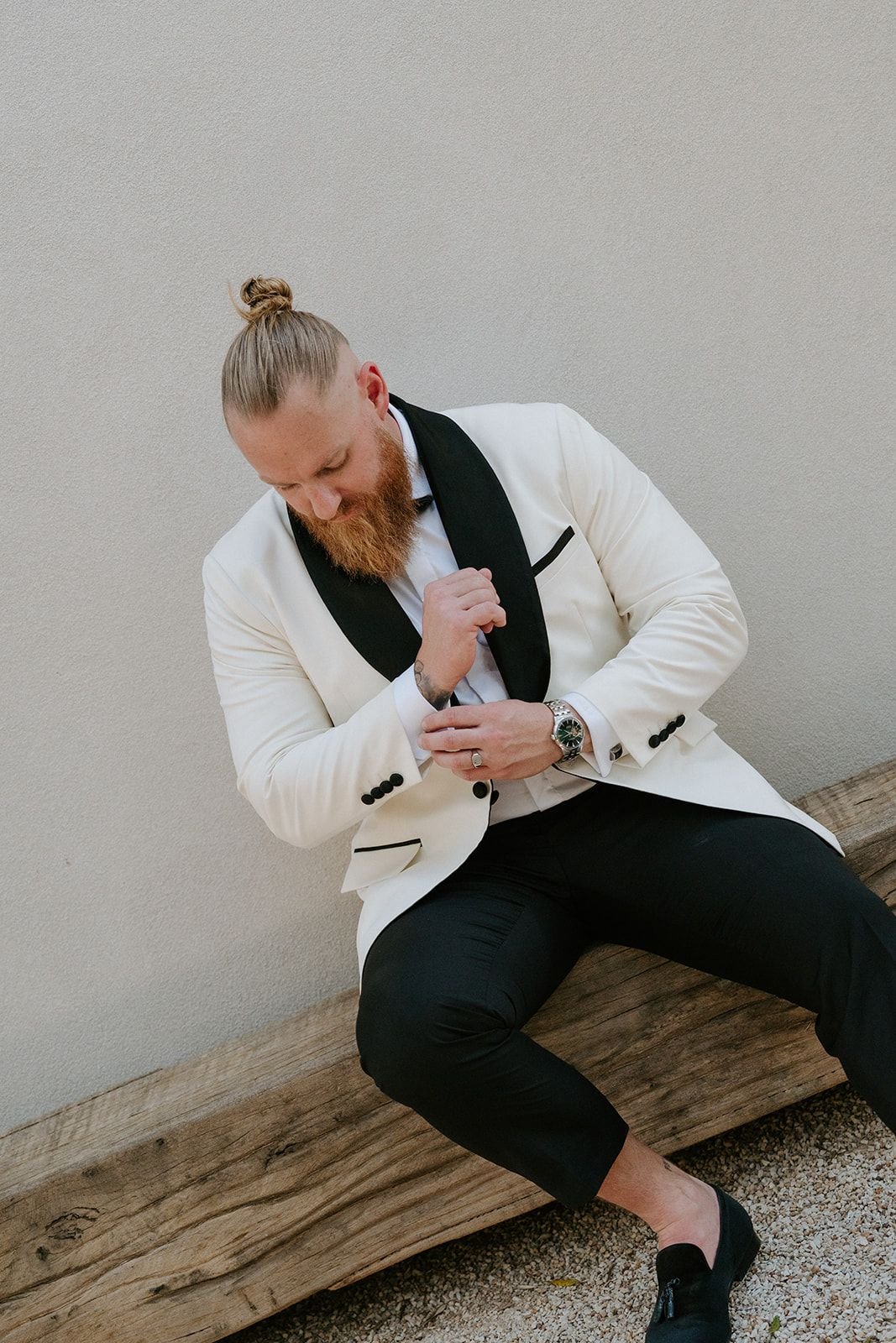 A man with a red beard and long hair tied in a bun, wearing a white tuxedo with black accents, sitting on a wooden bench against a plain wall, adjusting his sleeve.