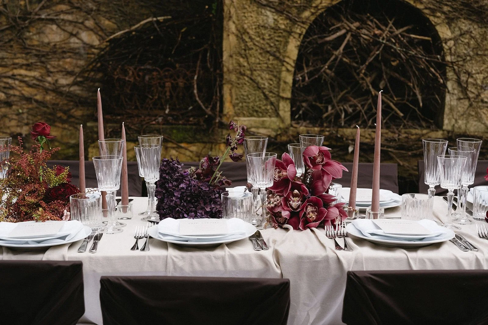Elegant table setting with white tablecloth, white plates, silverware, crystal glasses, pink candles, and purple and pink floral centerpieces, set against a stone wall background.