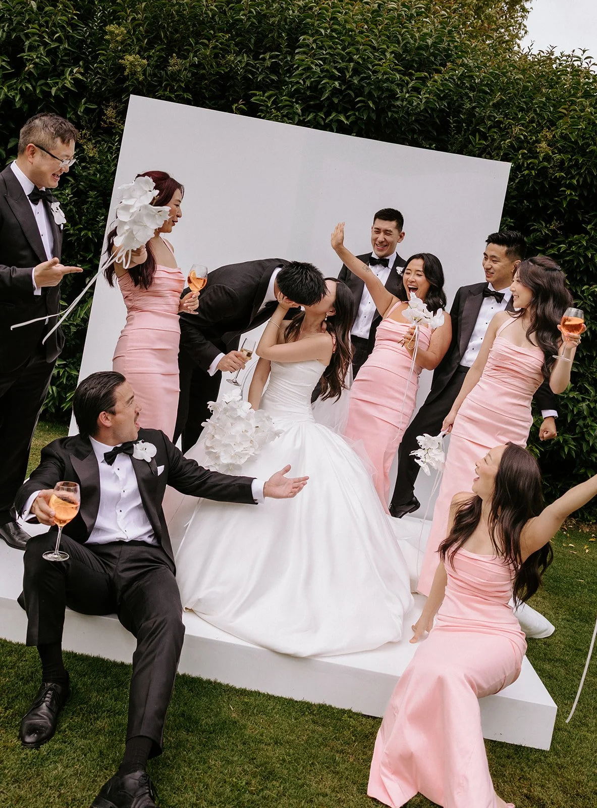 Group of wedding party members celebrating outdoors, with a bride in a white gown and bridesmaids in pink dresses, all smiling and holding glasses of rosé. The groom and groomsmen are dressed in black tuxedos, engaging joyfully.
