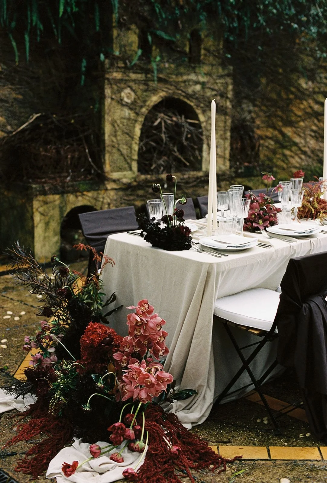 Elegant outdoor dining table decorated with flowers, tall candles, and glassware, set against a rustic stone wall with an arched opening surrounded by greenery.
