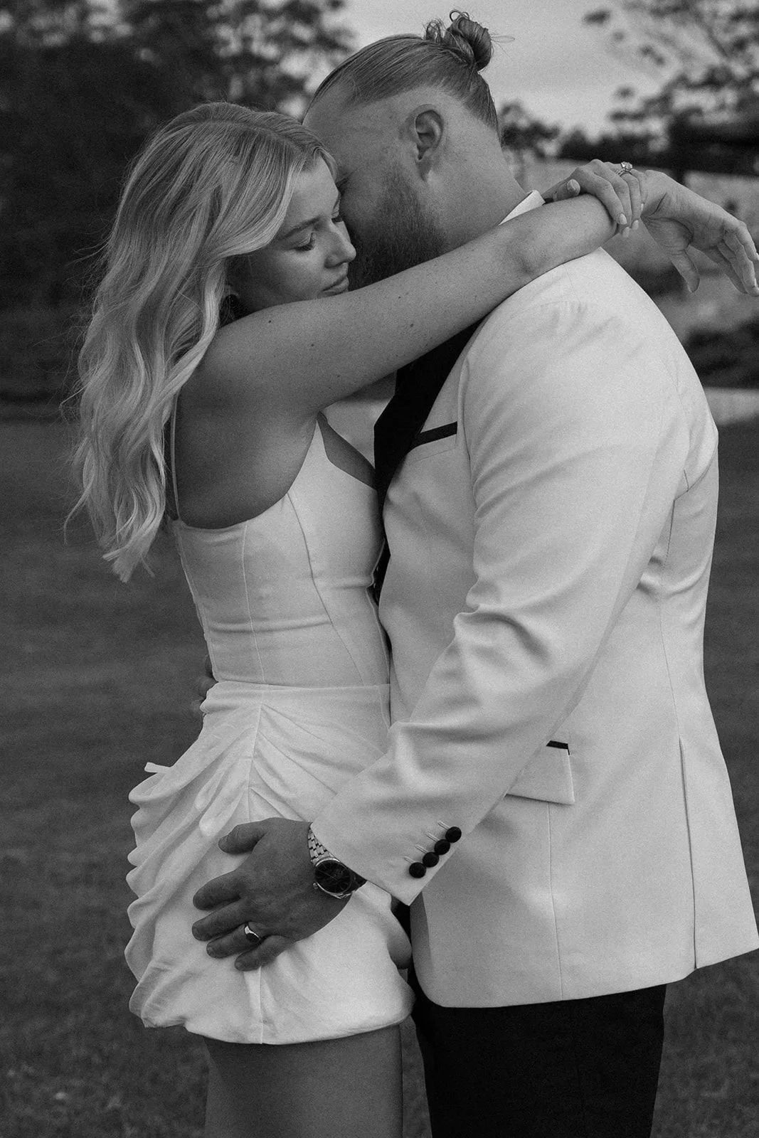 A black and white photo of a couple embracing outdoors, with their foreheads touching and eyes closed, in an intimate moment.