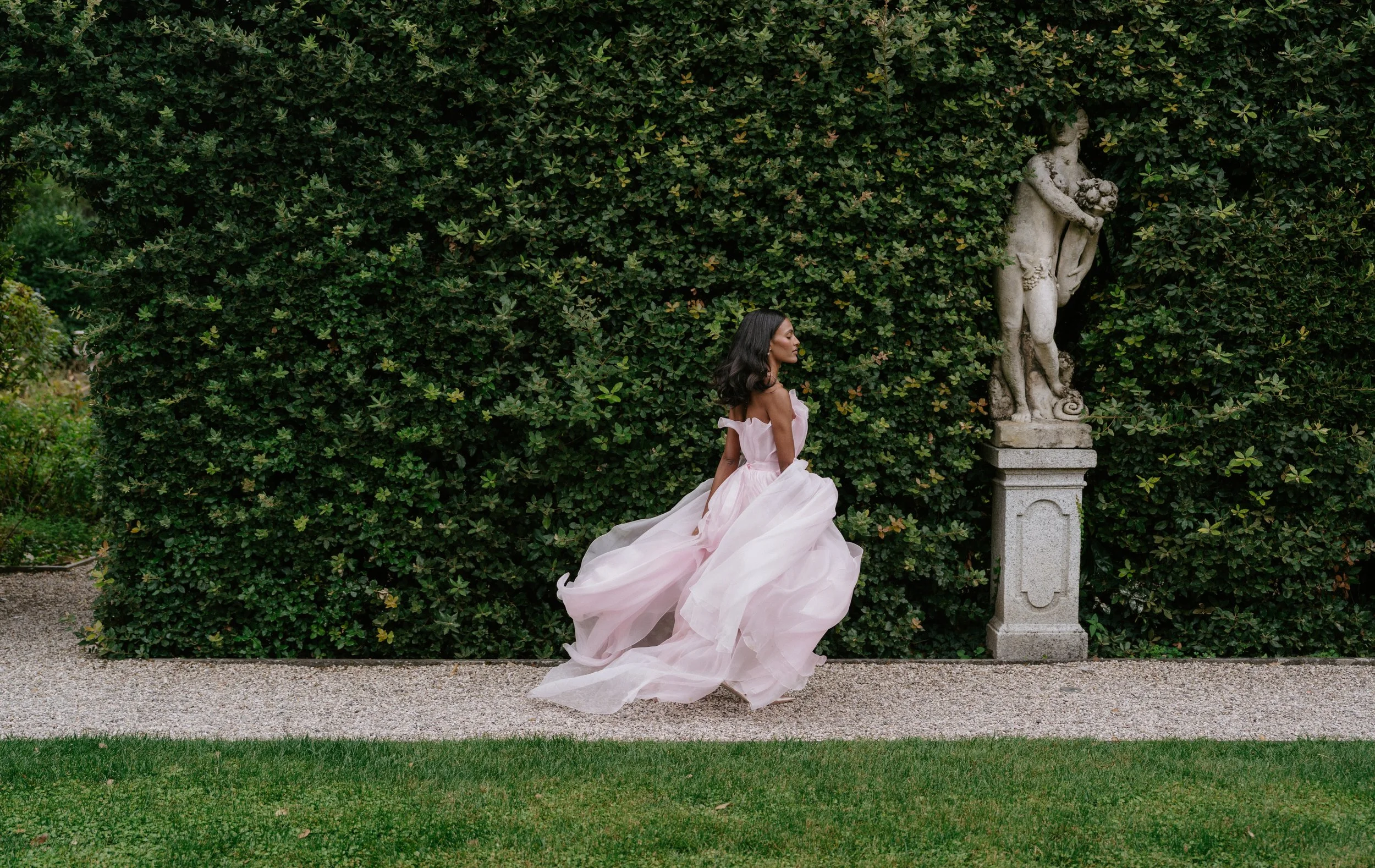 Brontë McVeigh wedding photography: A woman in a flowing pink gown walking past a stone sculpture of a male figure in a garden with lush green bushes
