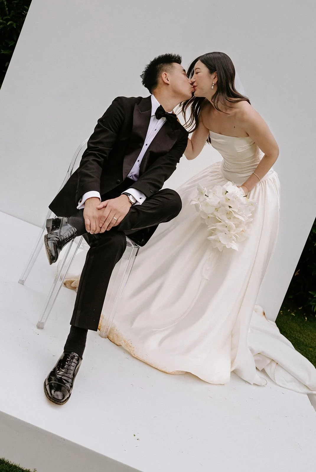 A bride and groom kissing during their wedding, with the bride holding a bouquet of white flowers and wearing a strapless satin wedding gown, while the groom is dressed in a black tuxedo and sitting on a clear acrylic chair.