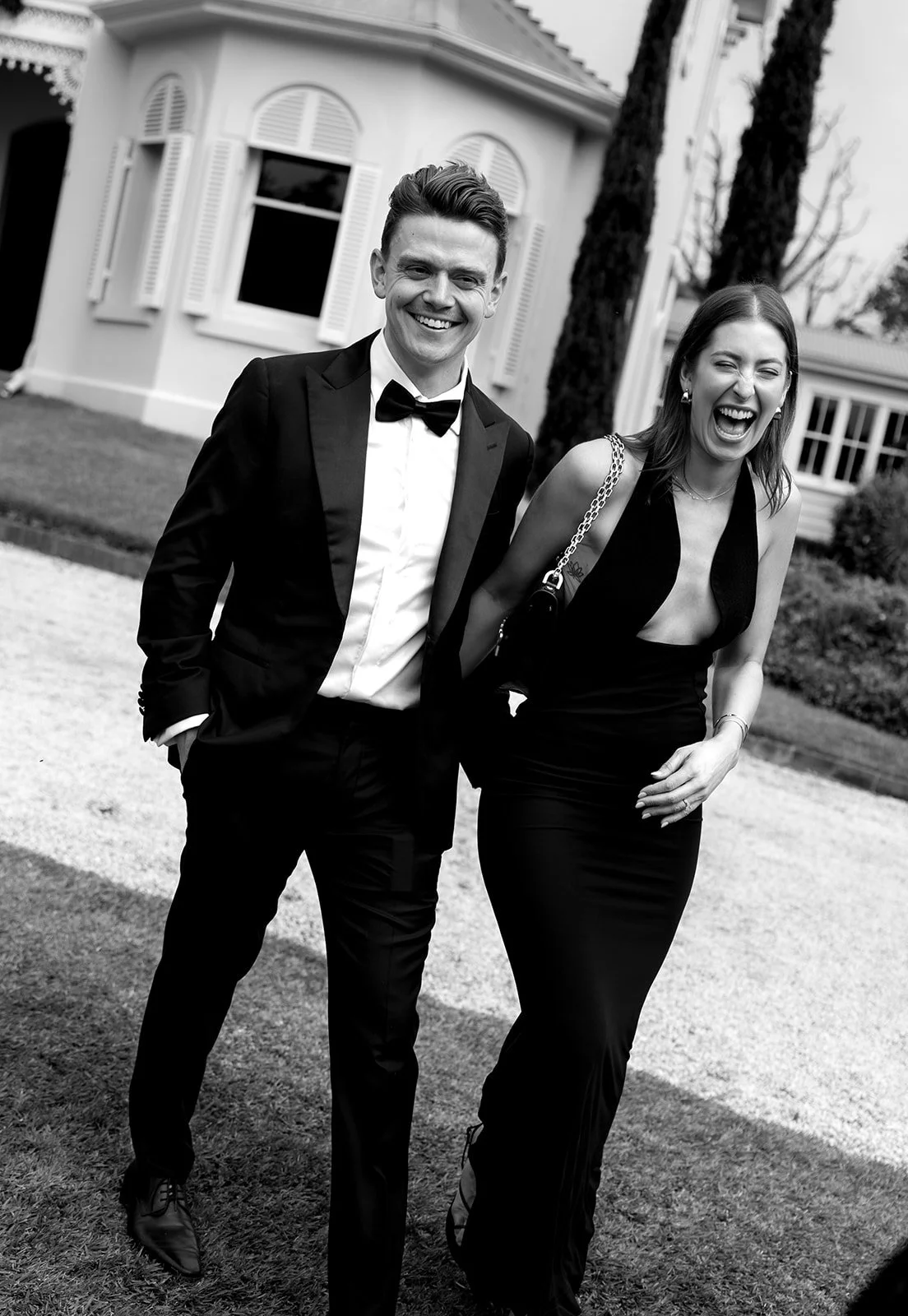 A man and woman dressed in formal attire smiling and laughing outdoors in front of a house with white walls and shuttered windows.
