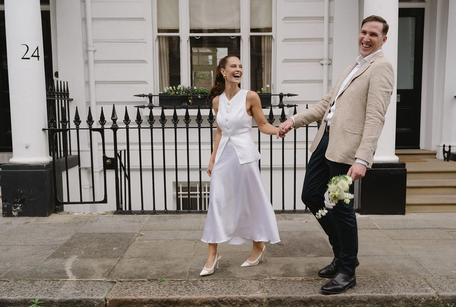 A couple dressed in wedding attire dancing and laughing on a sidewalk in front of a white building with black railings, the woman in a white dress and heels, the man in a beige jacket and dark pants holding a bouquet of white flowers.