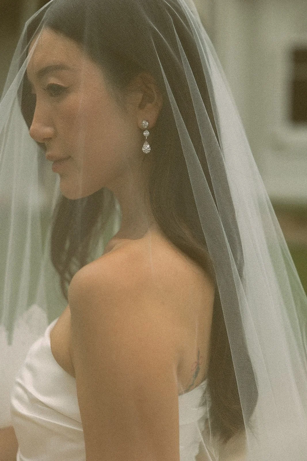 A bride with dark brown hair wearing diamond earrings and a white wedding dress, with a veil covering her head, looking to the side.