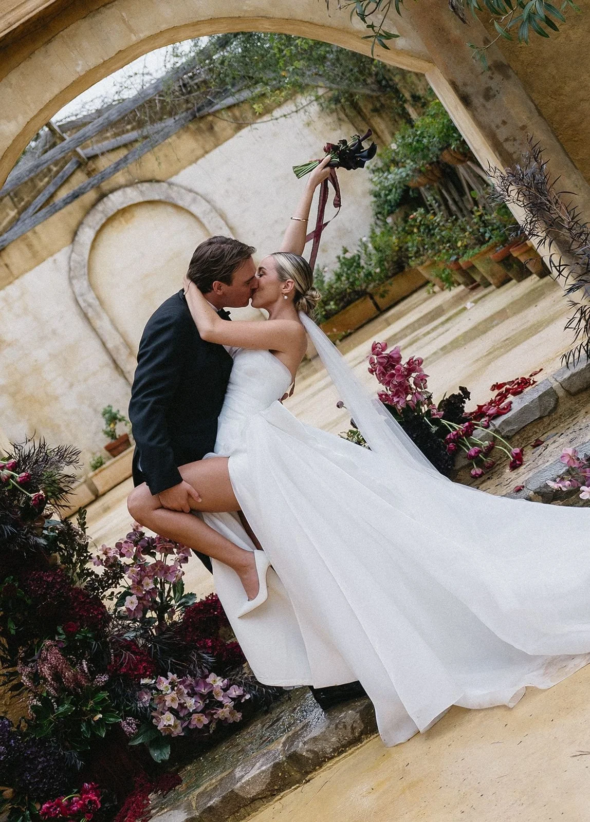 Wedding photography by Brontë McVeigh: A bride and groom sharing a kiss, with the bride lifting her right arm holding a bouquet, while the groom supports her, against a backdrop of an arched courtyard with plants and flowers.