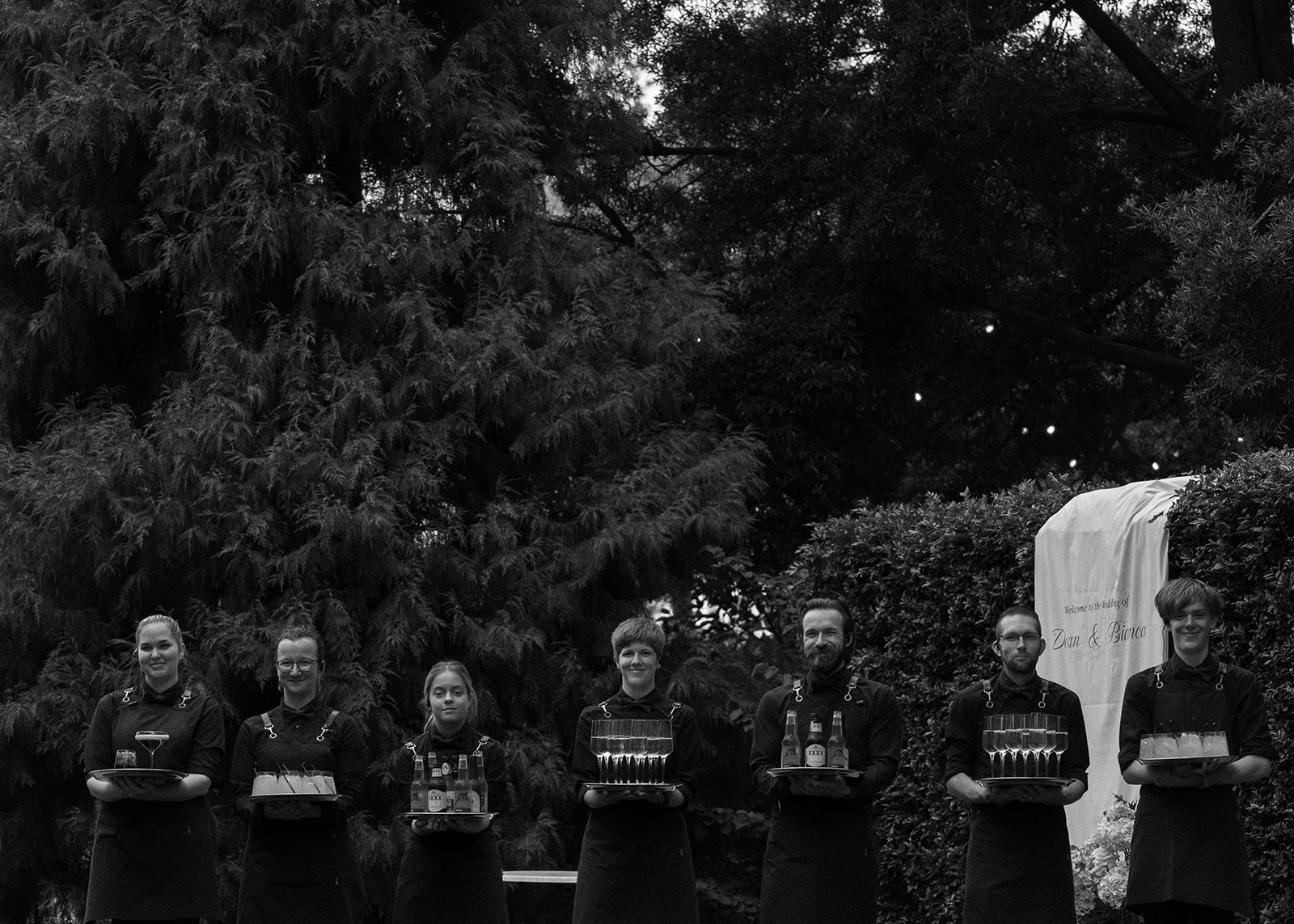 Seven servers standing outdoors in front of trees and shrubbery, holding trays of drinks, at a wedding reception for Dan and Bianca.
