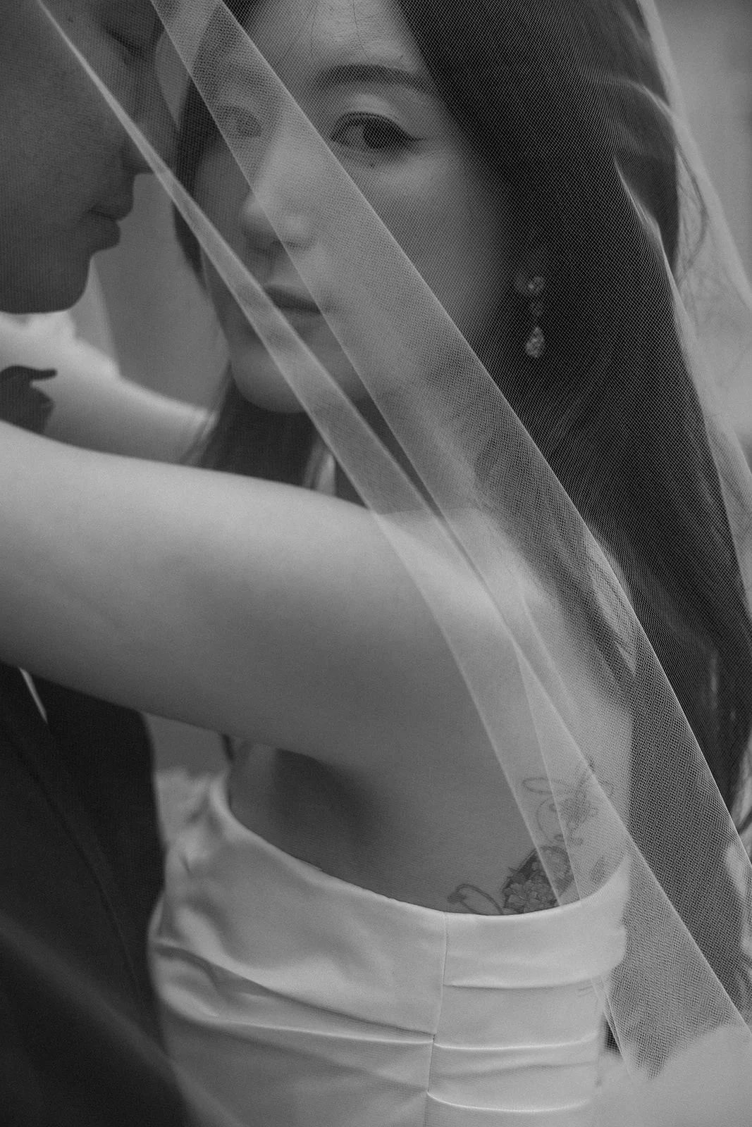 A black and white photo of two women, one with a veil covering her face, smiling softly, and the other close to her face, with her head slightly tilted.