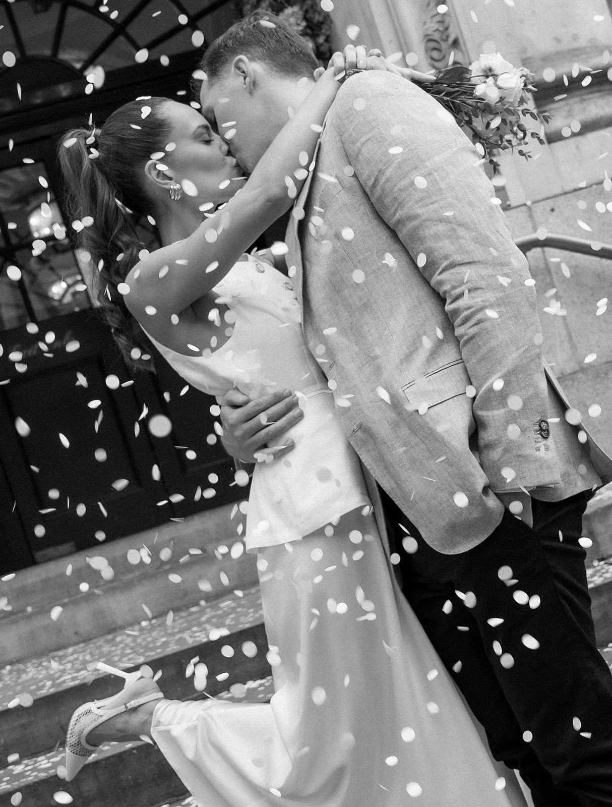 A couple in wedding attire hugging and kissing under falling confetti.