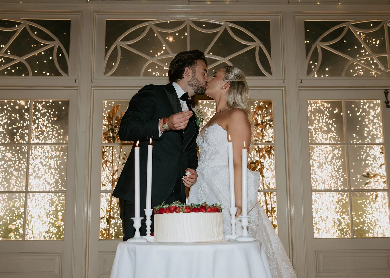 A bride and groom sharing a kiss during their wedding reception, standing behind a table with a wedding cake and tall candles, with dazzling sparklers in the background.
