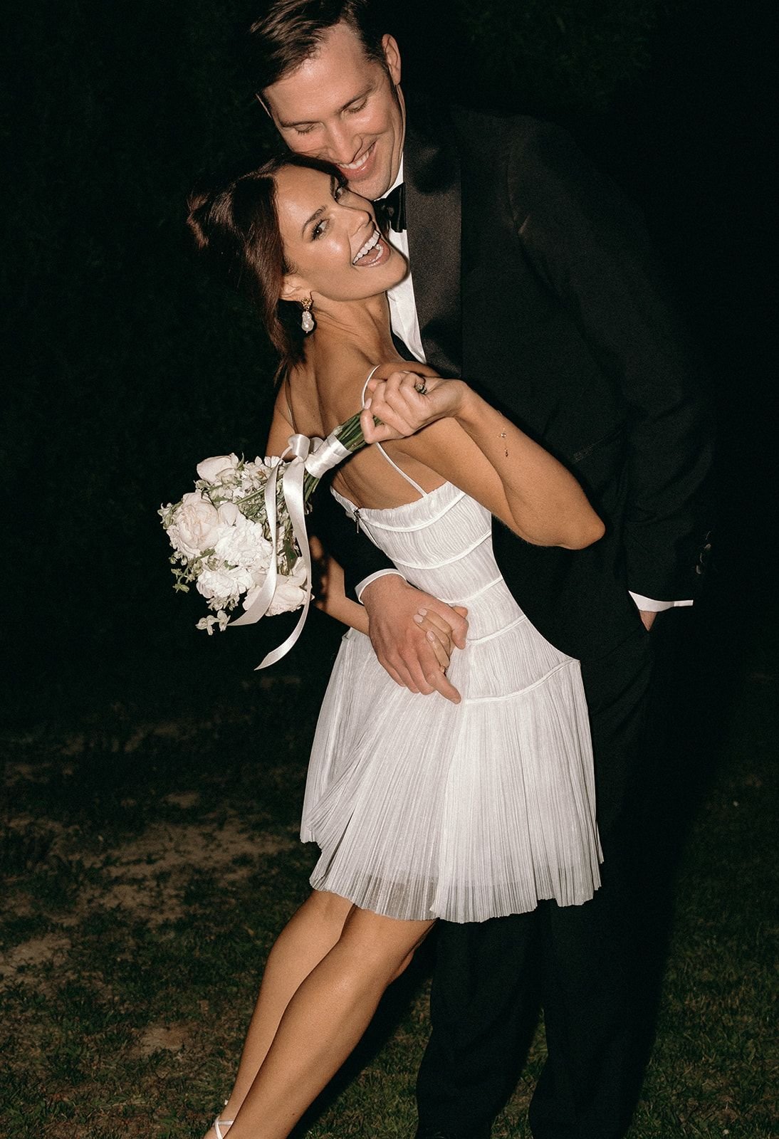 A couple dressed in formal attire, with the woman in a white dress holding a bouquet of white flowers, dancing and smiling at night.