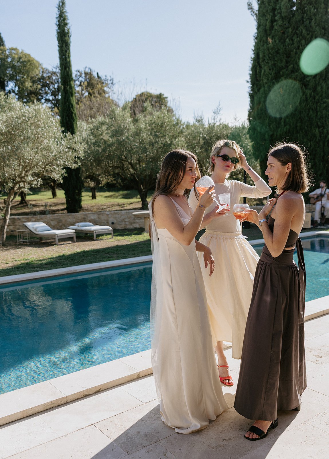 Three women in summer dresses standing by a swimming pool, holding cocktails and talking, with lounge chairs and trees in the background.