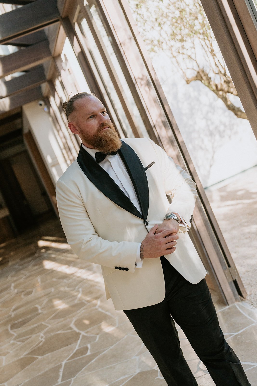 A man with a beard and slicked-back hair, wearing a white tuxedo jacket with black lapels, a white shirt, and a black bow tie, standing indoors near large windows with trees outside, looking thoughtful.