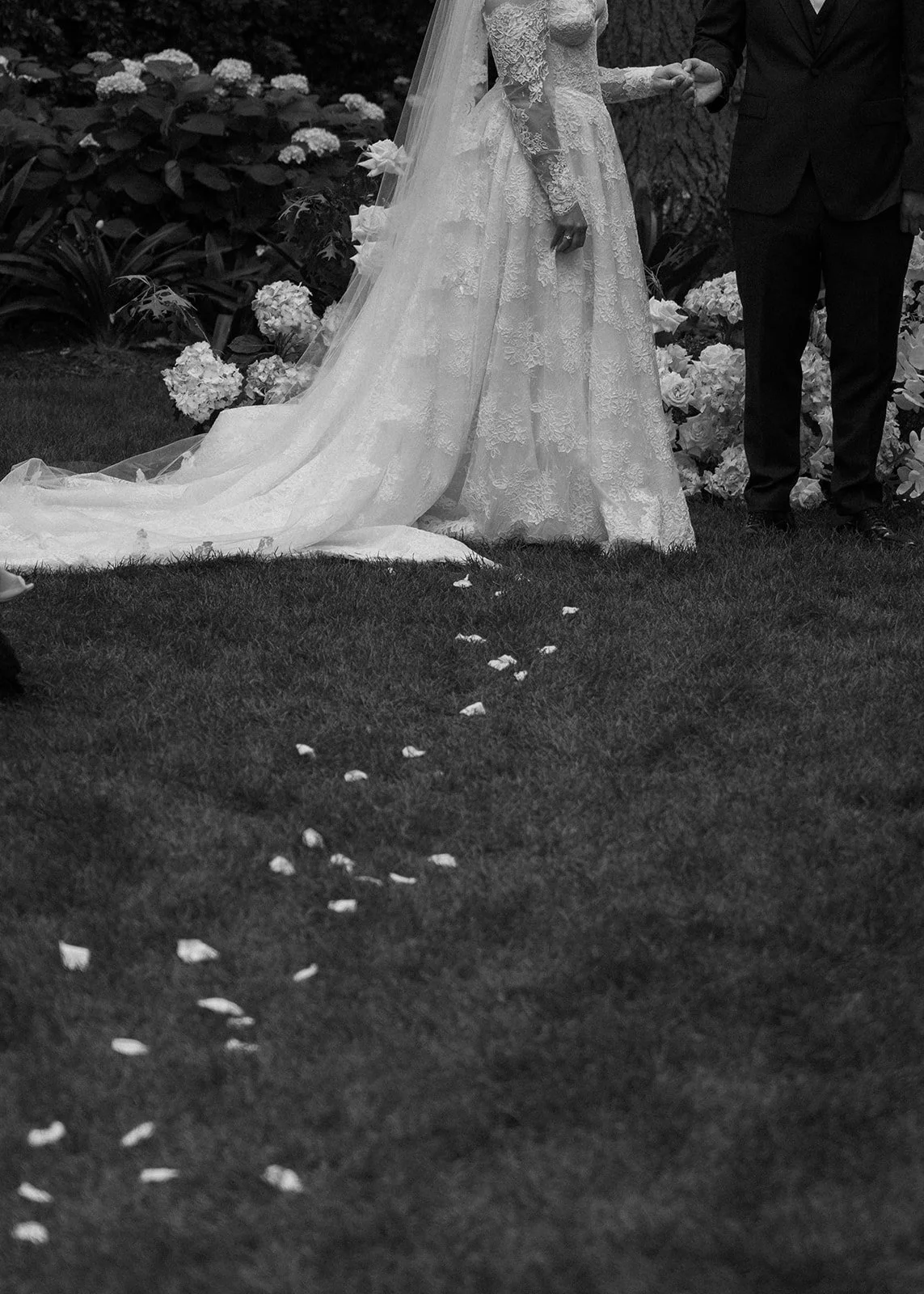 Black and white photo of a bride and groom holding hands during a wedding ceremony outdoors, with flower petals scattered on the grass.