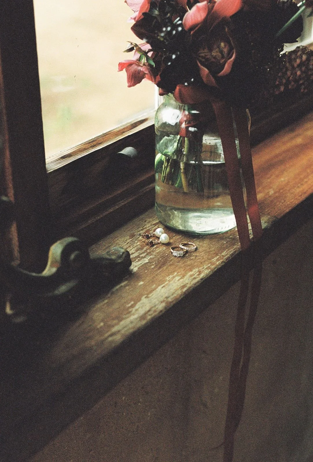 A wooden windowsill holding a glass vase with pink and dark flowers, some jewelry including pearl earrings and rings, near a window with a wooden frame.