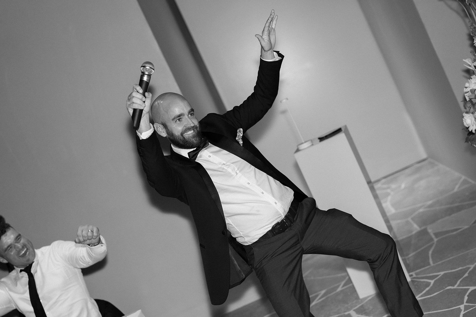 A man in a tuxedo holding a microphone and dancing at an event, smiling and raising his right hand, with another man in the background also smiling and acting cheerful.