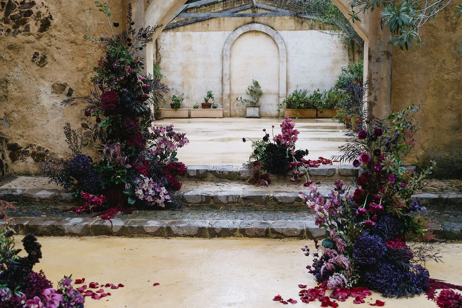 Decorated wedding or event altar with floral arrangements on stone steps, surrounded by stone walls and archways.