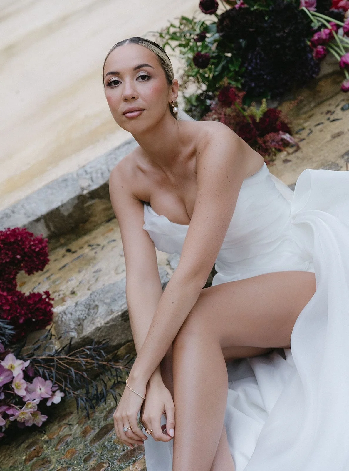A woman in a white dress is sitting outdoors on a stone surface, surrounded by colorful flowers and greenery.