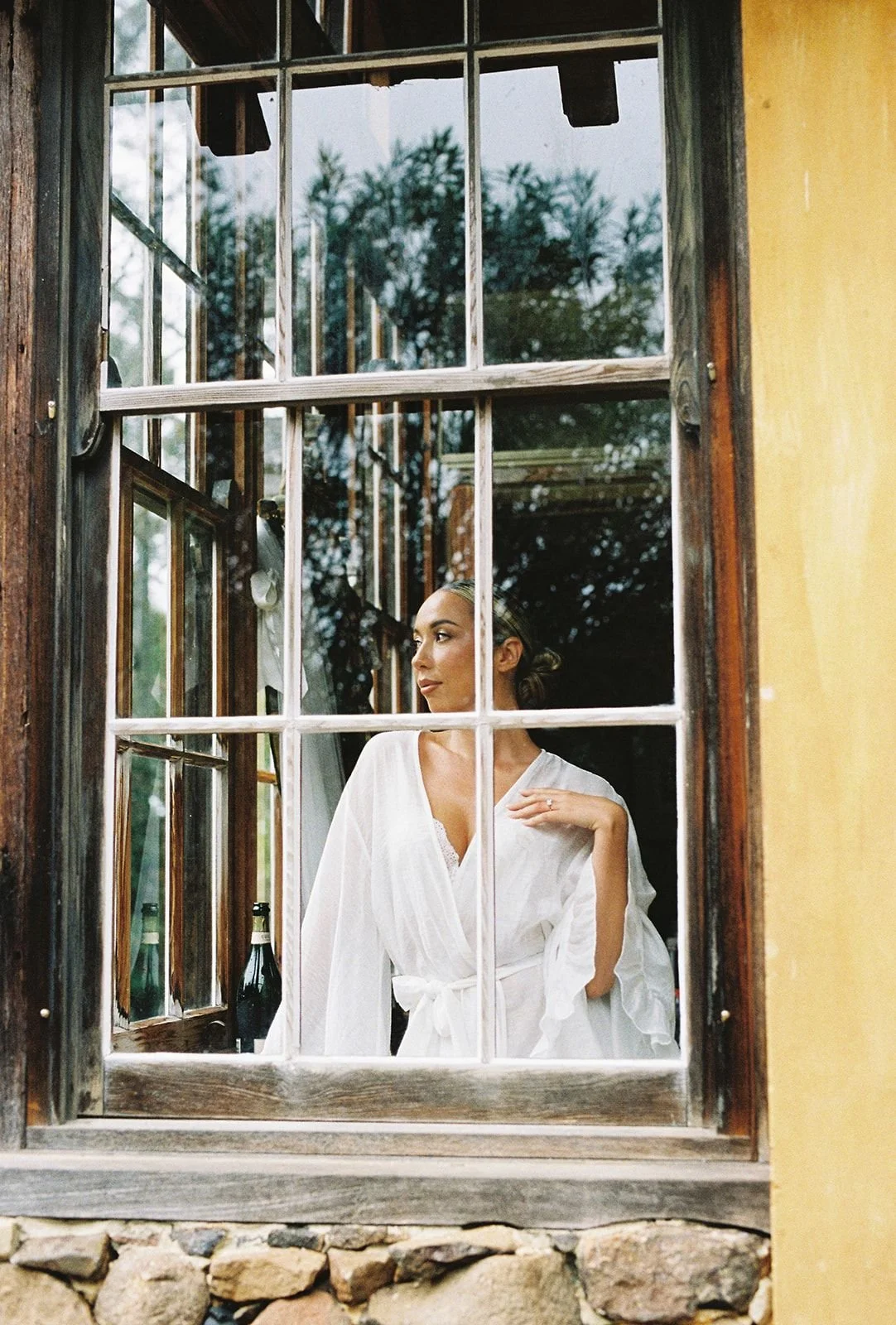 A woman in a white robe looking out a rustic wooden window with multiple panes, with bottles behind her and trees outside.