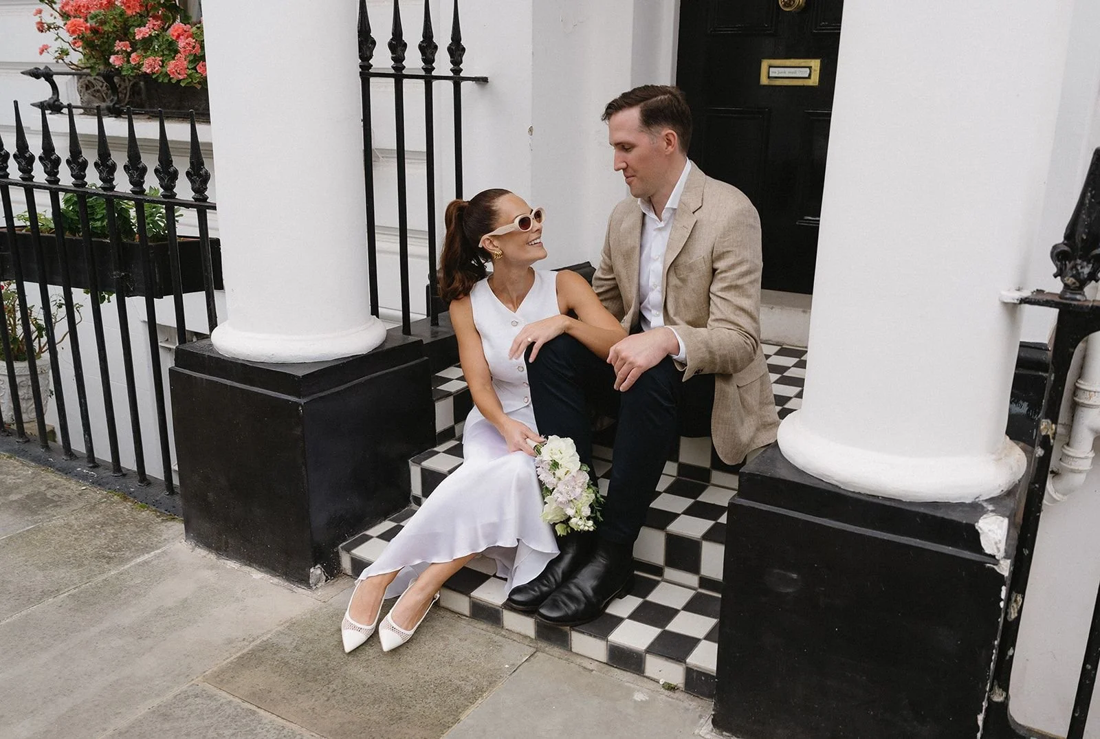 A woman in a white dress holding a bouquet sitting on the steps of a porch, smiling at a man in a beige blazer sitting nearby, both in front of white columns and a black door.