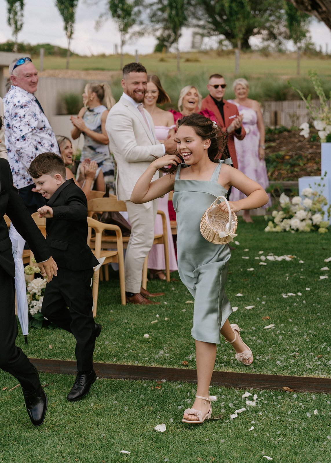 A young girl in a satin dress running with a basket at an outdoor wedding, smiling, as guests cheer and applaud in the background.