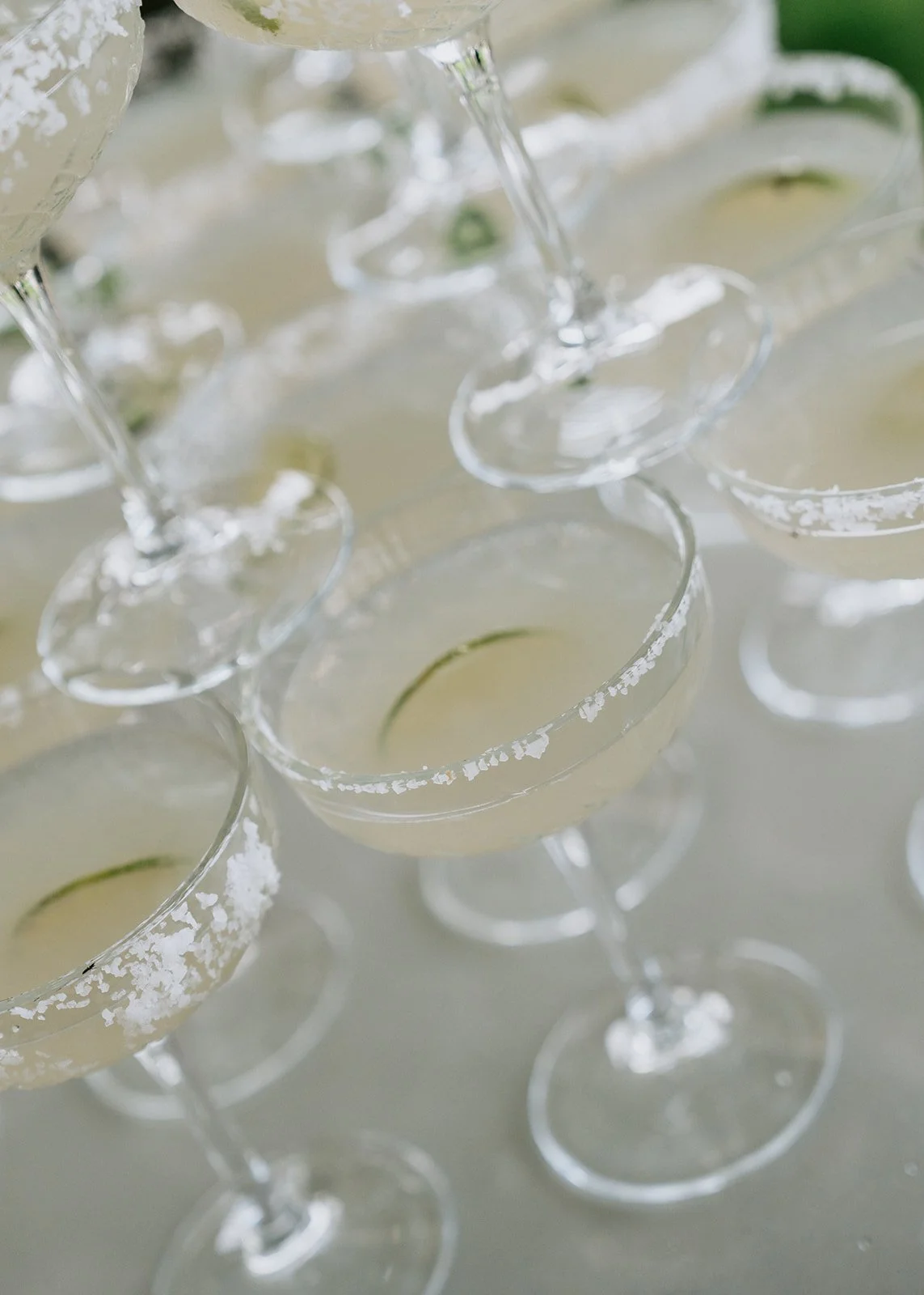 Several glasses of champagne garnished with lime slices, with salted rims, arranged on a white surface.