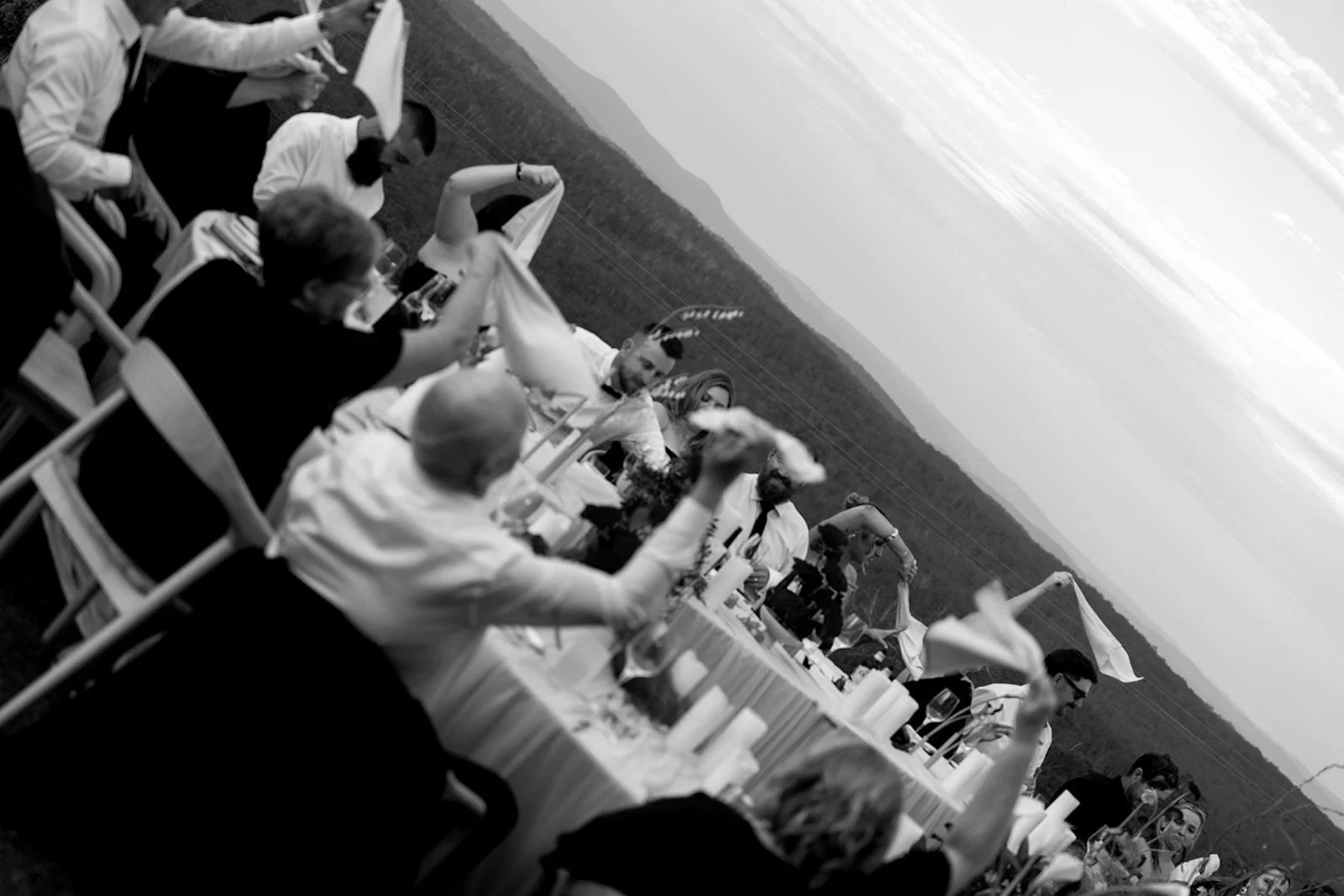 People seated at an outdoor dinner table with mountains and sky in the background, including some individuals holding flags, in black and white.