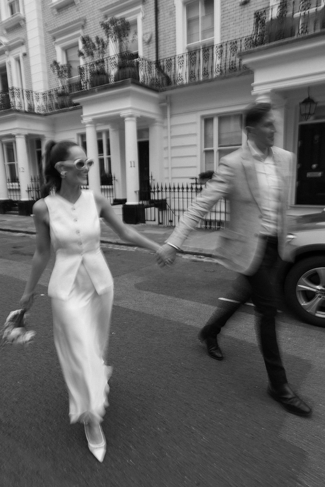 A black-and-white photo of a woman and a man holding hands while walking on a city street in front of white residential buildings with balconies.