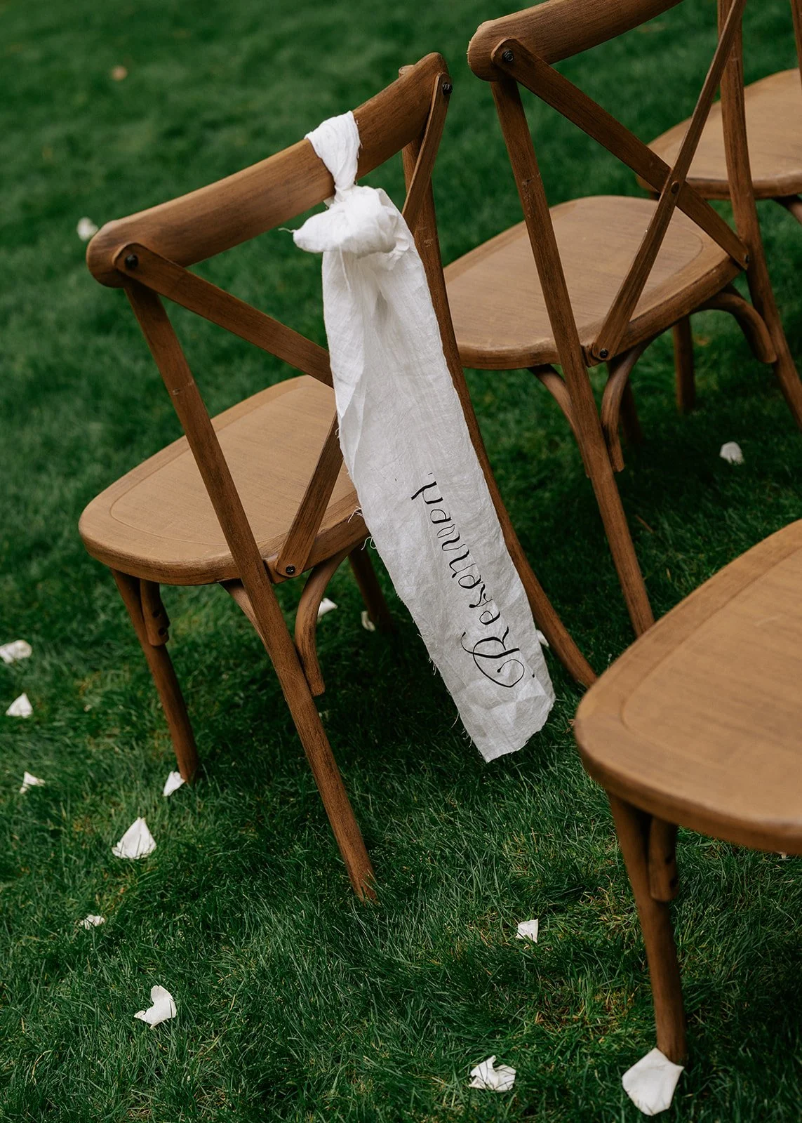 Wooden chairs on green grass with a white fabric sash reading 'Bride and' tied to the back of one chair, indicating a wedding ceremony setup.