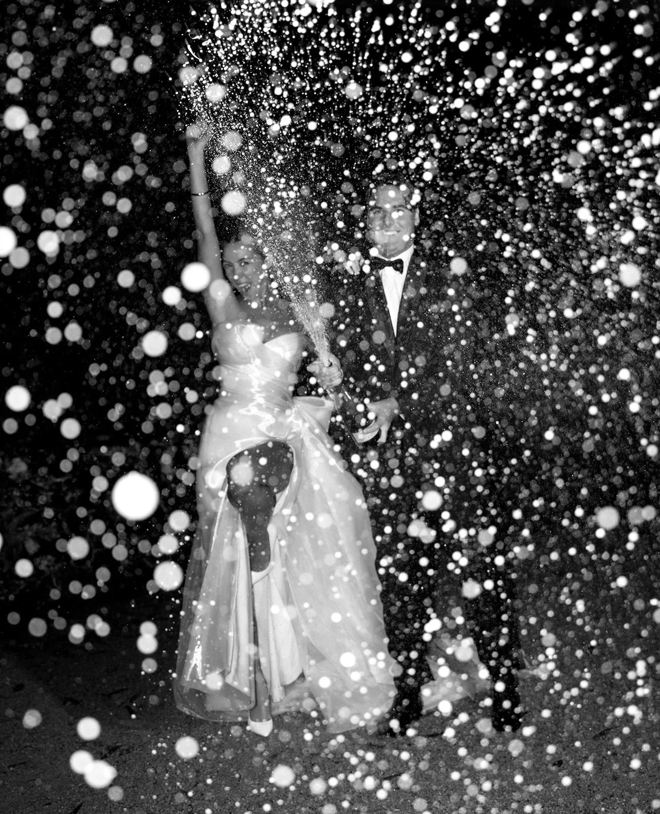 Black and white photo of a bride and groom celebrating with a champagne bottle and spray, surrounded by confetti at night.