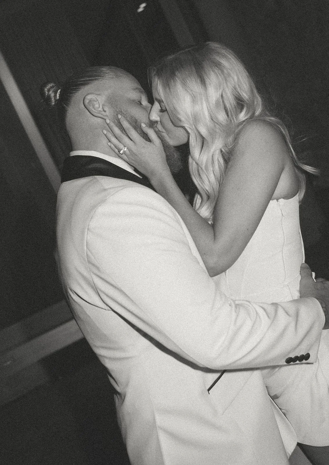 A black and white photo of a man and woman kissing, with the woman holding the man's face and wearing a ring, dressed in formal attire.