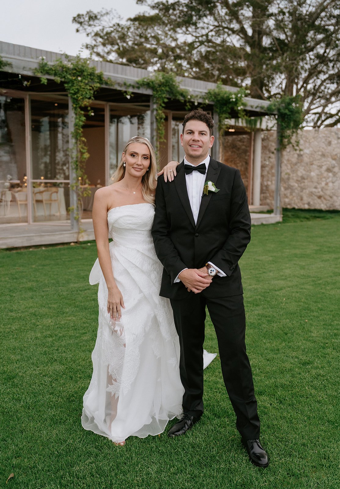 A bride and groom standing on a grassy lawn outdoors, with a modern glass building and trees in the background, dressed in wedding attire.