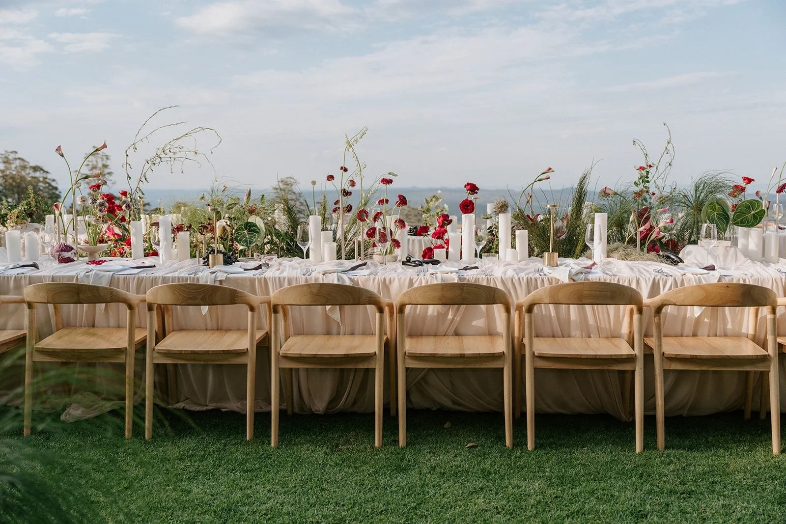 Elegant outdoor dining table decorated with floral arrangements, candles, and glassware, set against a scenic landscape with a body of water in the background.