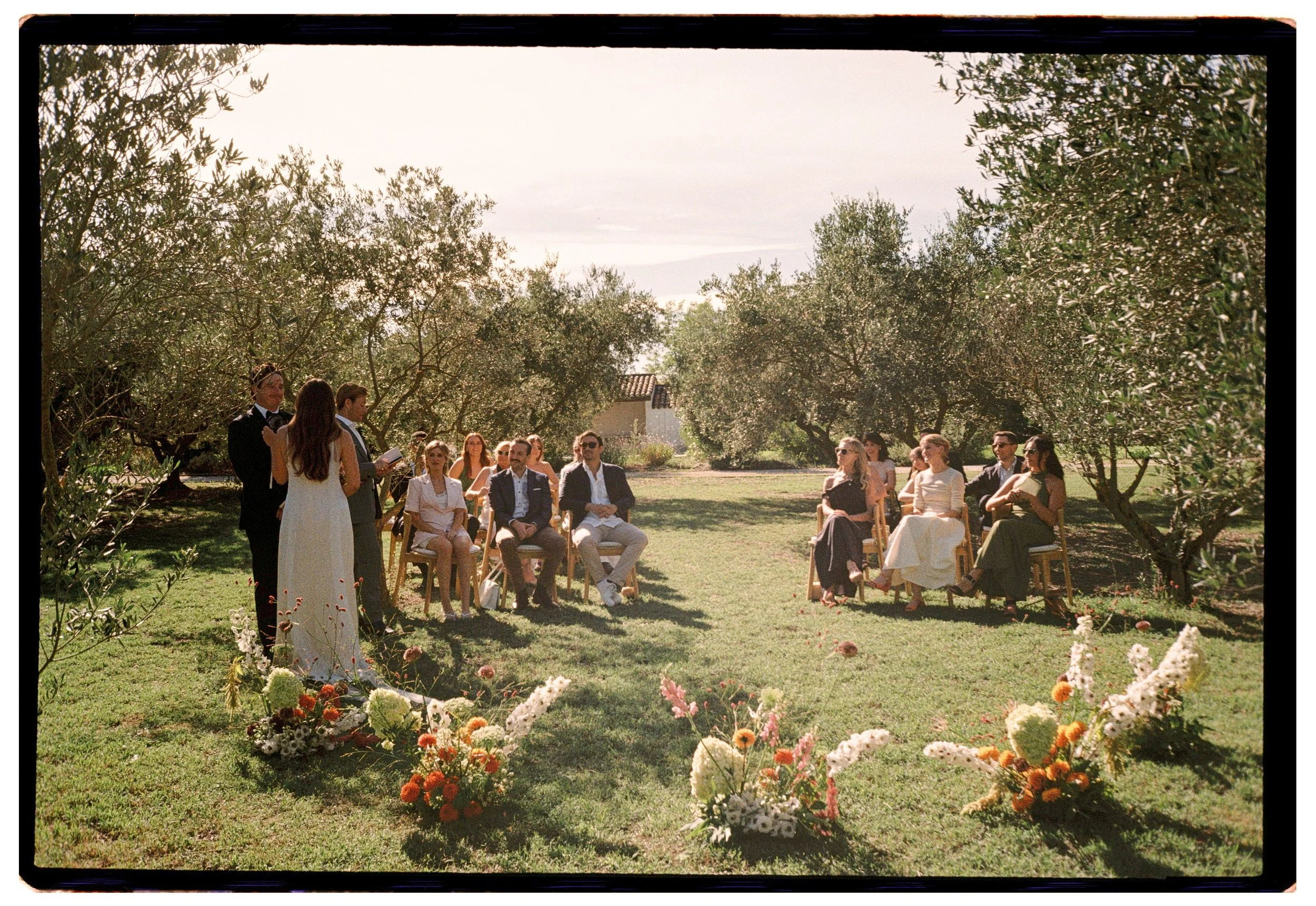 An outdoor wedding ceremony taking place in a garden with trees. The bride and groom are standing in front of seated guests, with floral arrangements on the grass near the ceremony area.